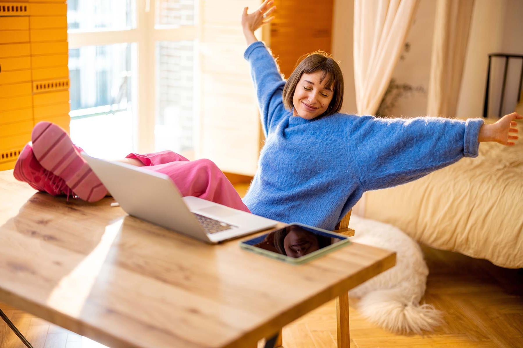 A woman stretching with joy at a table with a laptop.